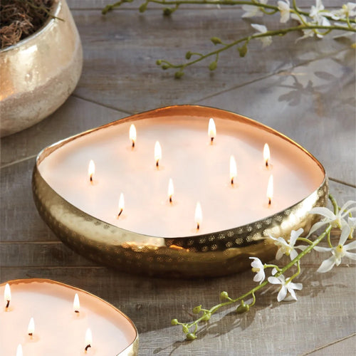 Oval gold bowl candle with multiple flames, placed on a wooden surface with white flowers nearby.