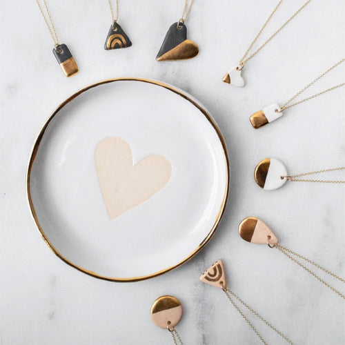 Gold and white ceramic dish with a heart, surrounded by geometric pendant necklaces on a marble surface.