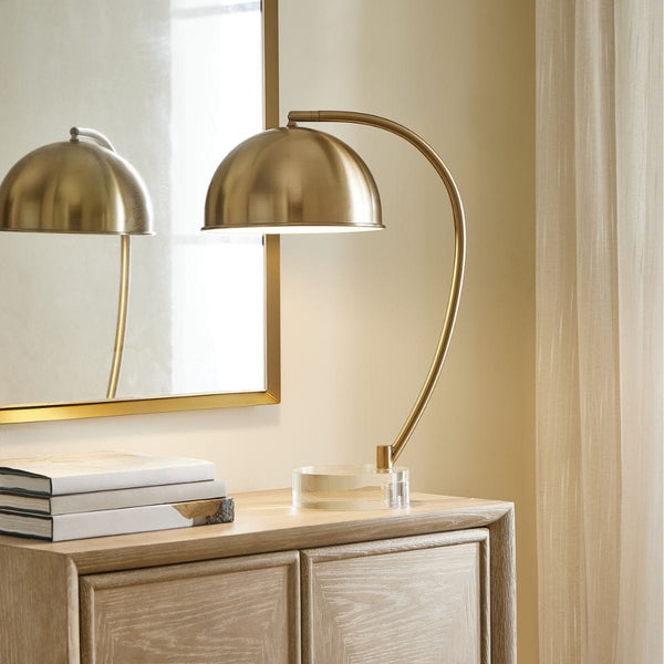 The Uttermost Romani Desk Lamp sits on a wooden cabinet beside stacked books, a mirror, and beige curtains.
