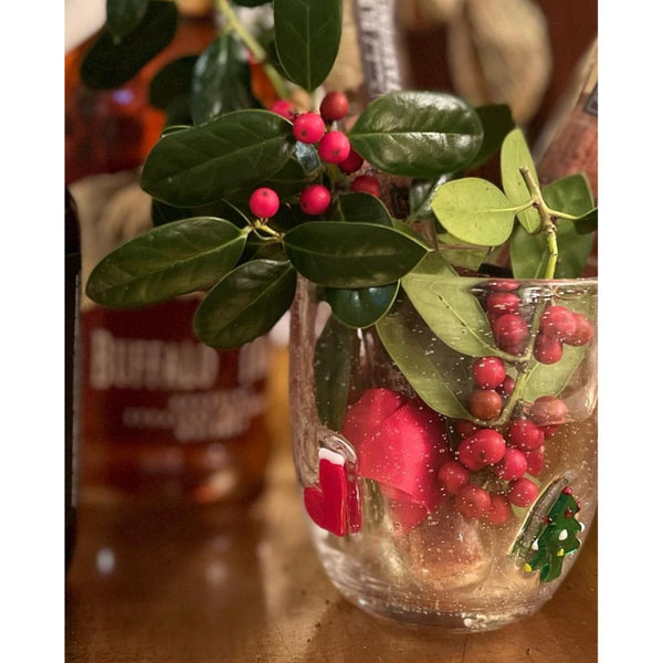 A set of Jingle Bubble Old-Fashioned Cocktail Glasses from Abigails, filled with holly branches, red berries, and other seasonal decorations, sits on a wooden surface. Bottles are in the background. These exquisite glasses are handmade in Italy, adding an extra touch of elegance to any holiday display.