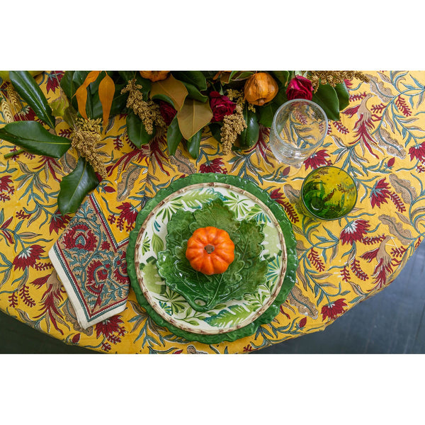 Decorative table setting with a green plate, small pumpkin, and glass on a yellow floral tablecloth.