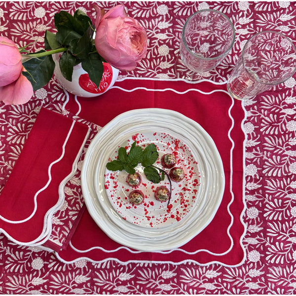 Table setting with a plate of food, glasses, and a pink flower on a red patterned tablecloth.