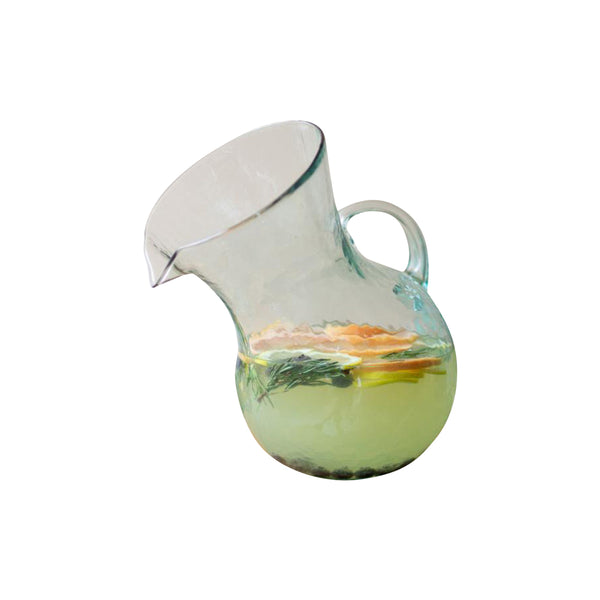 The Kalalou Tilted Clear Glass Cocktail Pitcher, crafted from mouth-blown recycled materials, is perfect for serving refreshing water filled with lemon and herbs.