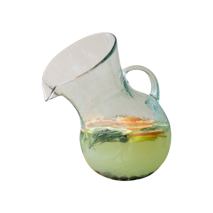 The Tilted Clear Glass Cocktail Pitcher by Kalalou is a unique, mouth-blown pitcher crafted from recycled materials, designed to hold a lemon and herb infused beverage in its handle.