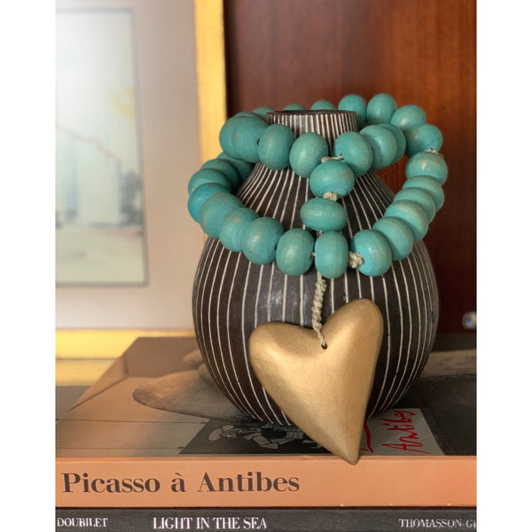 Creative Co-Op Turquoise + Gold Beaded Heart Strand decorates art books, including Picasso, atop a mango wood vase.