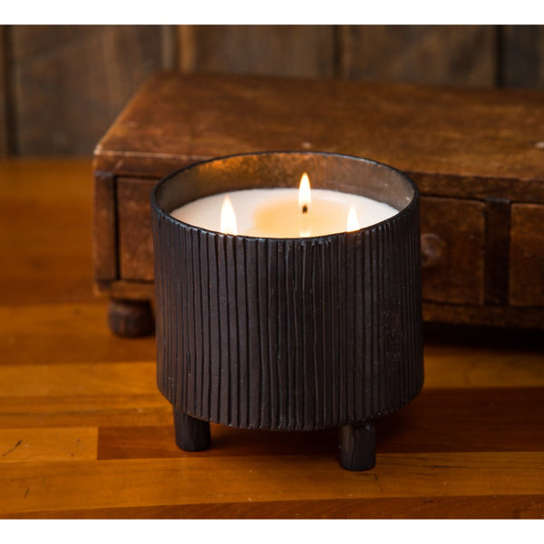 The Himalayan Trading Post Blacksmith Forged Candle in a ribbed black vessel sits on wood, with a rustic wooden box behind.