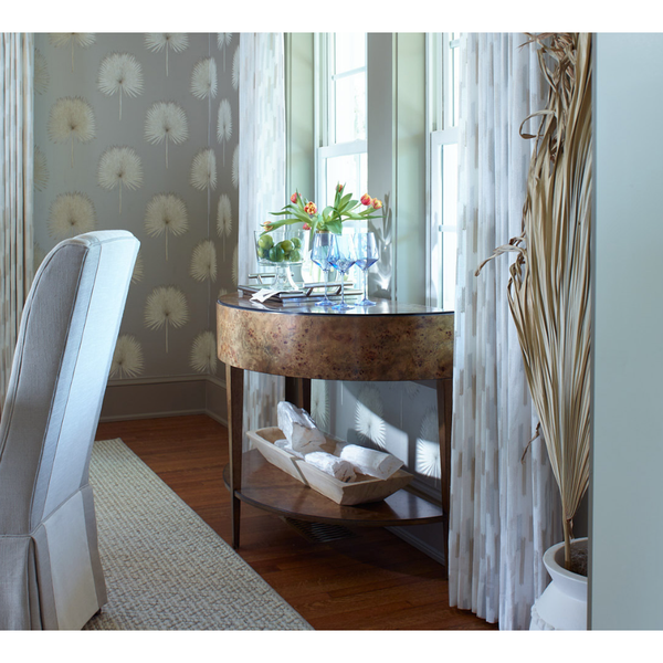 The Isla Natural Burl Demilune Console by Woodbridge Furniture sits by tall windows with glassware, flowers, and pillows.