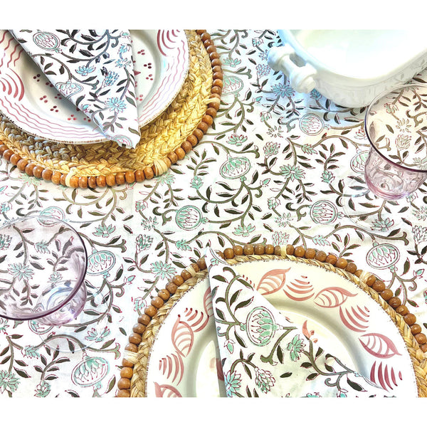 The table is set with Majolica ceramics, folded napkins, woven placemats, and pink glasses on a floral tablecloth. Casa Nuno Pink Dinner Plates by Abigails add a touch of elegance to the arrangement.