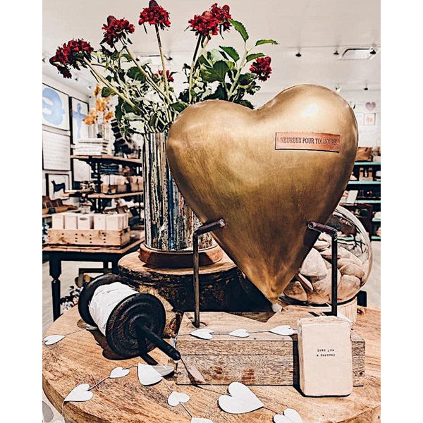 The Heureux Pour Toujours Unique Metal Heart on Wooden Base by Sugarboo & Co. is displayed on a wooden table alongside red flowers in a vase and love-themed decor, creating an ideal anniversary gift shop setting.