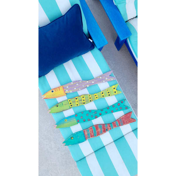 Hand-painted, vibrant fish sculptures crafted from reclaimed distressed wood by Kalalou rest on a striped blue and white lounge chair, accompanied by a cushion and another chair. These colorful Painted Recycled Wood Wall Art pieces (set of 4) bring joy to the serene setting.