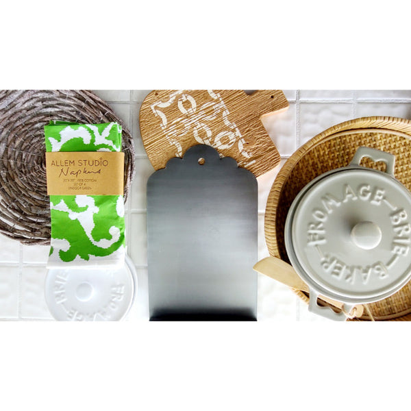 Kitchen items on a table include a green napkin, wooden cheese board, metal cutter, and a stainless steel cheese baker set in a woven tray.