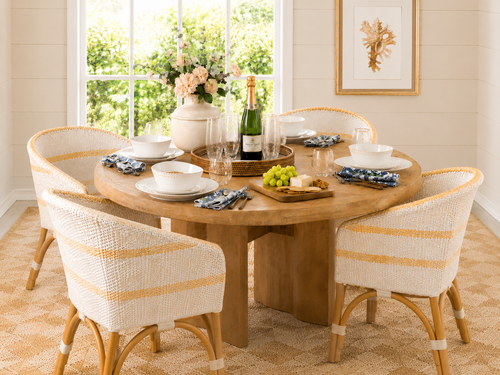 A round wooden dining table set for four with white chairs, plates, glasses, a vase of flowers, champagne, and a tray of fruit, near a bright window and framed wall art.