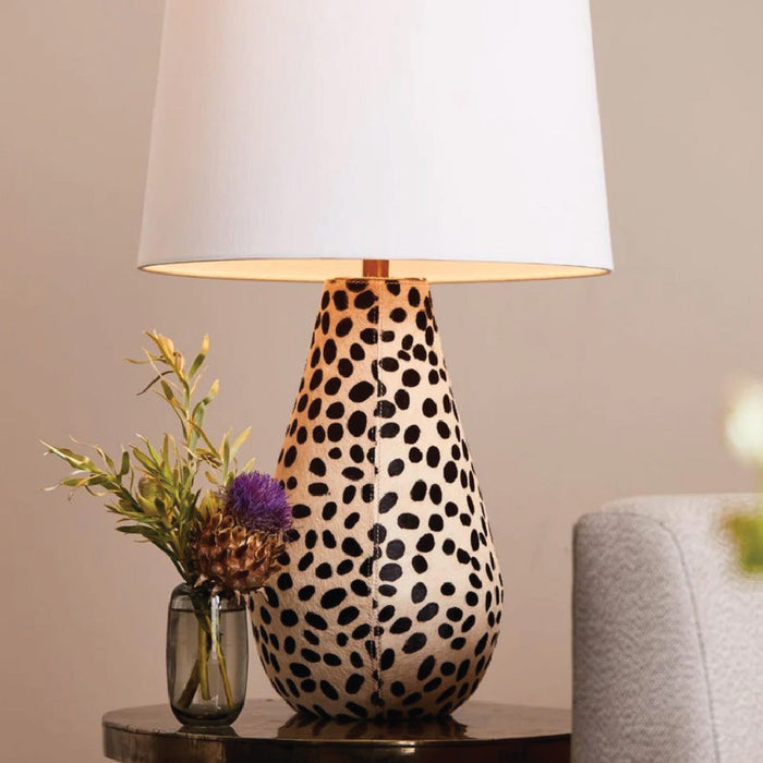 Table lamp with a white shade and a black spotted base, next to a small vase with flowers on a dark table.