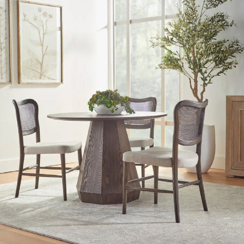 A round wooden dining table with four lattice-back chairs, set on a beige rug, against a green hedge wall.