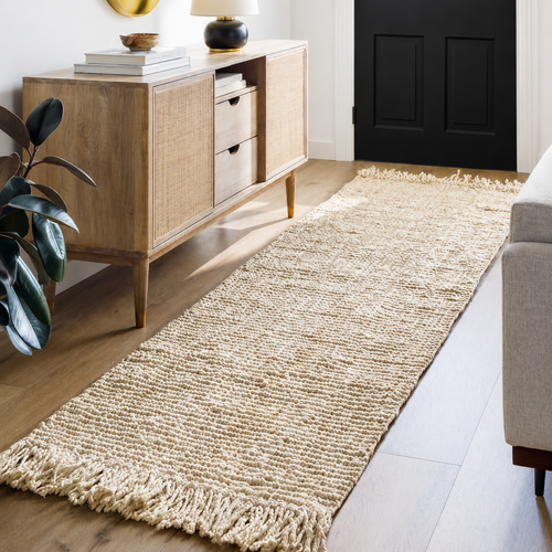 A beige woven rug with fringe on a wooden floor beside a sideboard and plant in a bright entryway.