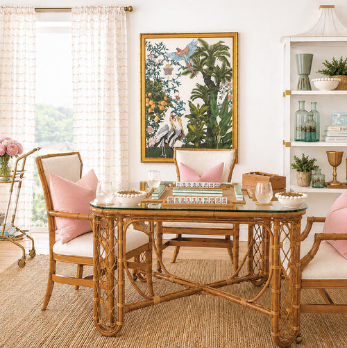 A bright dining room with a bamboo table set for mahjong, pink-cushioned chairs, tropical art on the wall, a bar cart, and plants, all on a jute rug by sunny windows with sheer curtains.