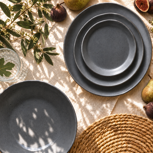 A table setting with stacked gray plates, figs, pears, a woven placemat, a glass of water with a leaf, and olive branches on a cream tablecloth in natural sunlight.