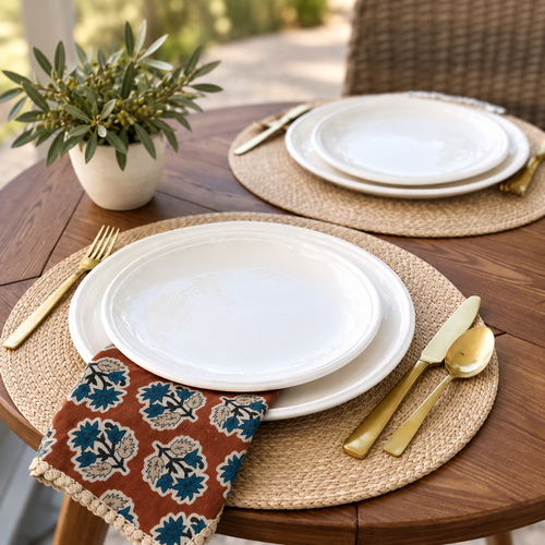 Two place settings on a wooden table, each with white plates, gold cutlery, and a woven placemat, with a small potted plant in the center.