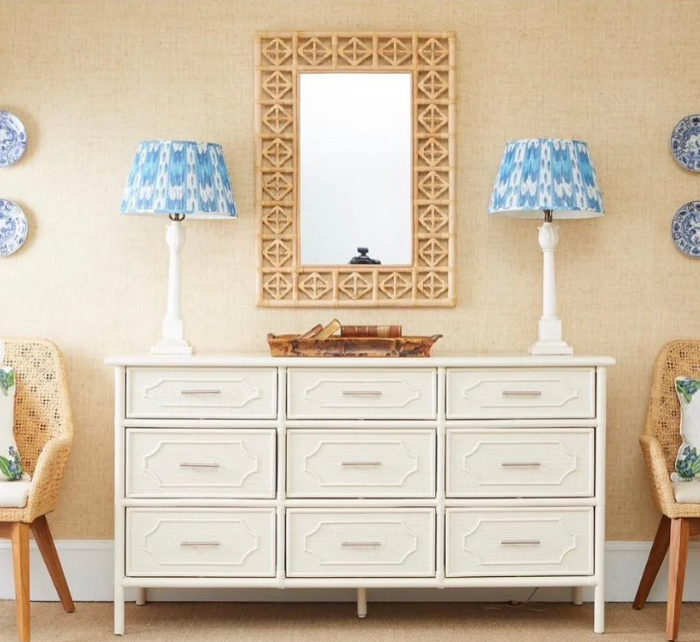 A pink wavy-edged mirror above a white table with a lamp, books, and a vase with white flowers.