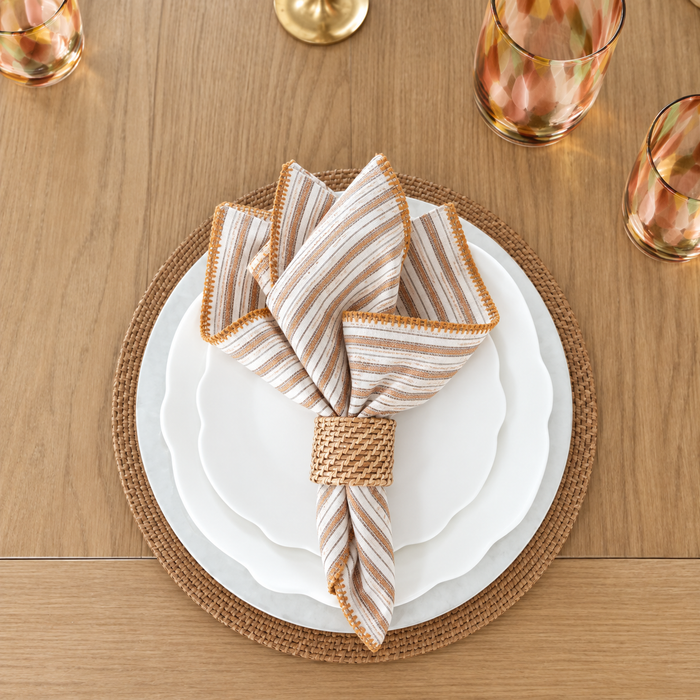 A striped cloth napkin in a woven ring sits on stacked white plates with a wicker placemat, surrounded by patterned tumblers on a wooden table.