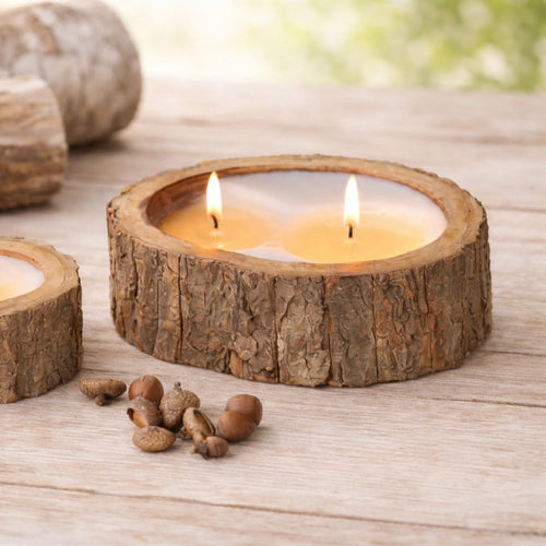 Two lit candles in rustic wooden holders sit on a wooden table with scattered acorns nearby.