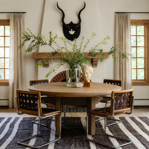 Round wooden table with four chairs, greenery in vases, and décor in a room with large windows and a fireplace.