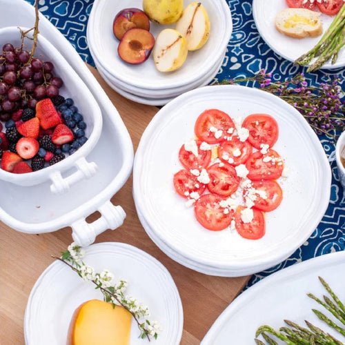 White dishes with sliced tomatoes, fruit, berries, cheese, and asparagus on a blue patterned tablecloth.