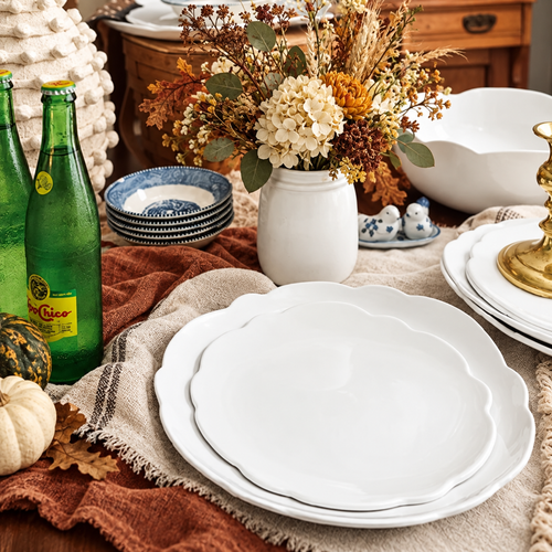 A table setting featuring stacked white scalloped plates, green glass bottles, and a seasonal floral centerpiece.