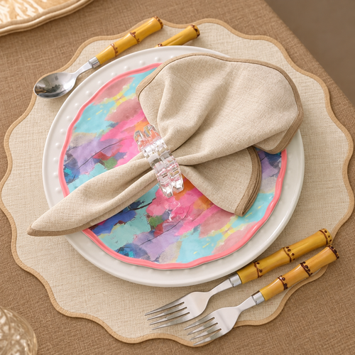 A place setting with a white plate, a watercolor-patterned accent plate, a beige napkin in a clear ring, and bamboo-handled silverware.