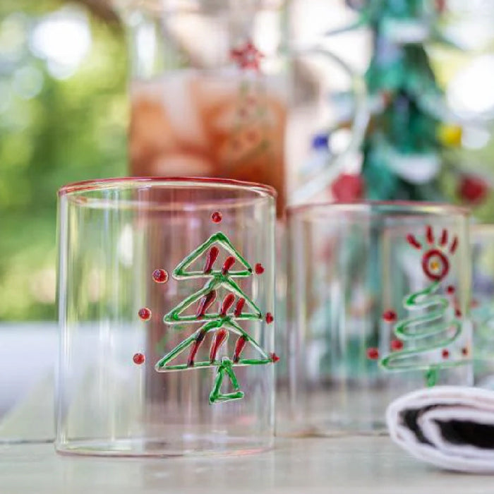 Clear glass with a painted Christmas tree and red dots, with another decorated glass blurred in the background.