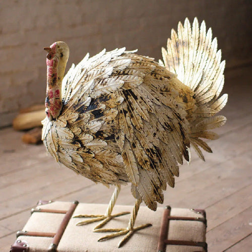 A decorative metal turkey sculpture with detailed, gold-toned feathers stands on a trunk indoors.