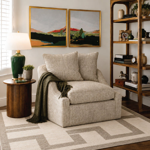 A cozy beige armchair with green throw blanket, near a side table, bookshelf, and landscape paintings.