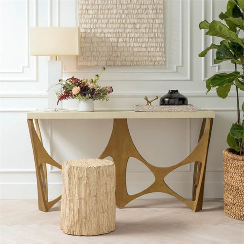Modern console table with gold geometric legs, a stone stool, a lamp, flowers, and potted plant beside it.