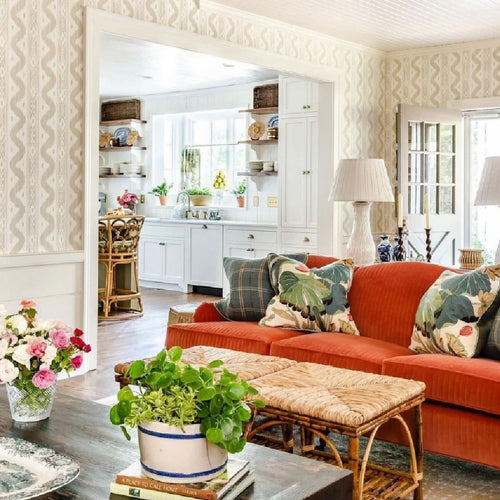 Bright living room with an orange sofa, floral pillows, plants, and a view into a white kitchen.