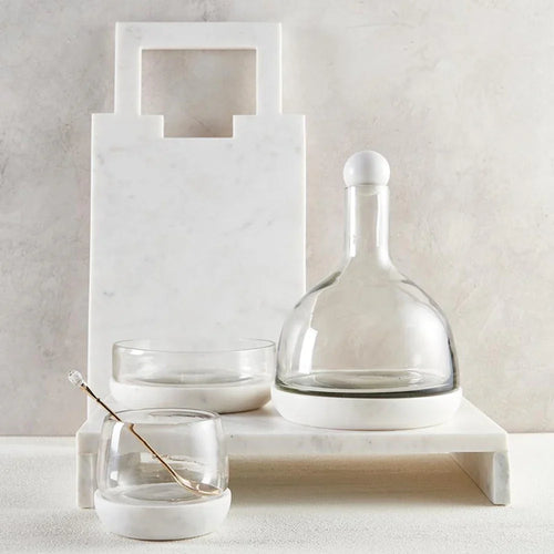 Marble cutting board, glass decanter, and bowls with a decorative spoon on a white surfaced background.