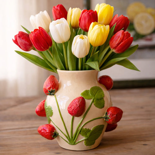 A ceramic vase with strawberries holds red, yellow, and white tulips on a wooden table.
