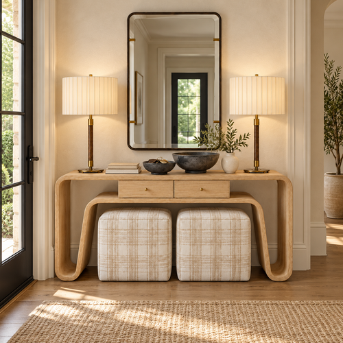 A cozy living room with a brown rug, wooden bench, bookshelf, console table with lamps, artwork, and decorative accents in warm neutral tones. Large window lets in natural light.