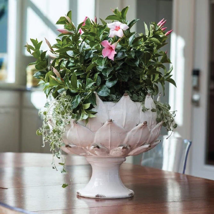 A white ceramic pedestal planter with pink flowers and green foliage sits on a wooden table indoors.
