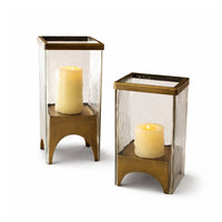 Two brass candle holders with clear glass covers containing lit candles on a white background