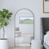 The Uttermost Crosley Oversized Mirror reflects light beside a bed, plant, and ottoman in a bright bedroom.