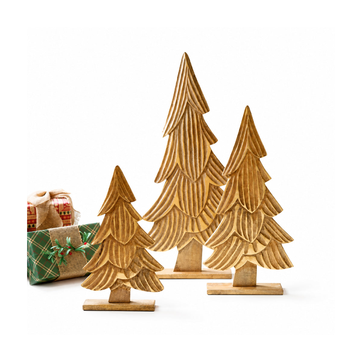 Three small wooden Christmas tree ornaments with a layered, scalloped texture, arranged in a size progression from left to right on a white background, with two wrapped gifts partially visible behind the leftmost tree.