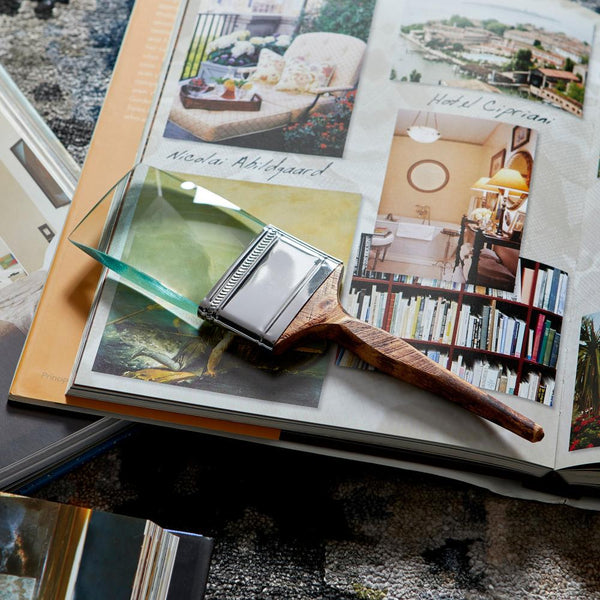 A wooden-handled paintbrush with a silver metal ferrule rests across an open art book or magazine. The book pages show interior design and landscape photographs, and the brush is placed on a translucent green card over a textured dark surface.