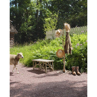 A rustic outdoor scene with a wooden coat rack holding three straw hats and a brown coat, beside a small bench and boots on a gravel path. A sheep walks in the background among green plants, a white picket fence, and trees.