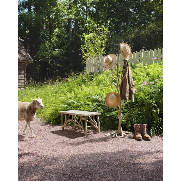 A rustic outdoor scene with a wooden coat rack holding three straw hats and a brown coat, beside a small bench and boots on a gravel path. A sheep walks in the background among green plants, a white picket fence, and trees.