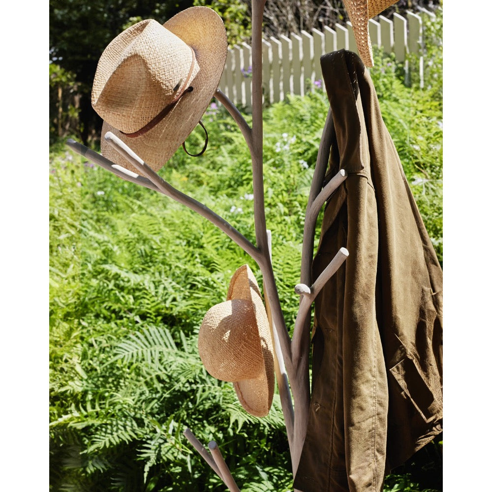 A rustic branch-style coat rack outdoors, holding a wide-brim straw hat, a folded olive-brown jacket, and another straw hat, with green foliage and a white picket fence in the background.