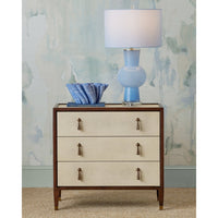 Three-drawer dresser with a blue lamp and decorative bowl against a patterned wall.