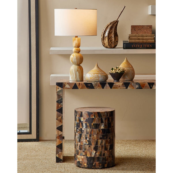 A decorative console table with a geometric mosaic shell-patterned base and white floating shelves, styled in a warm interior setting with a table lamp, vases, books, and a matching shell-covered cylindrical stool.