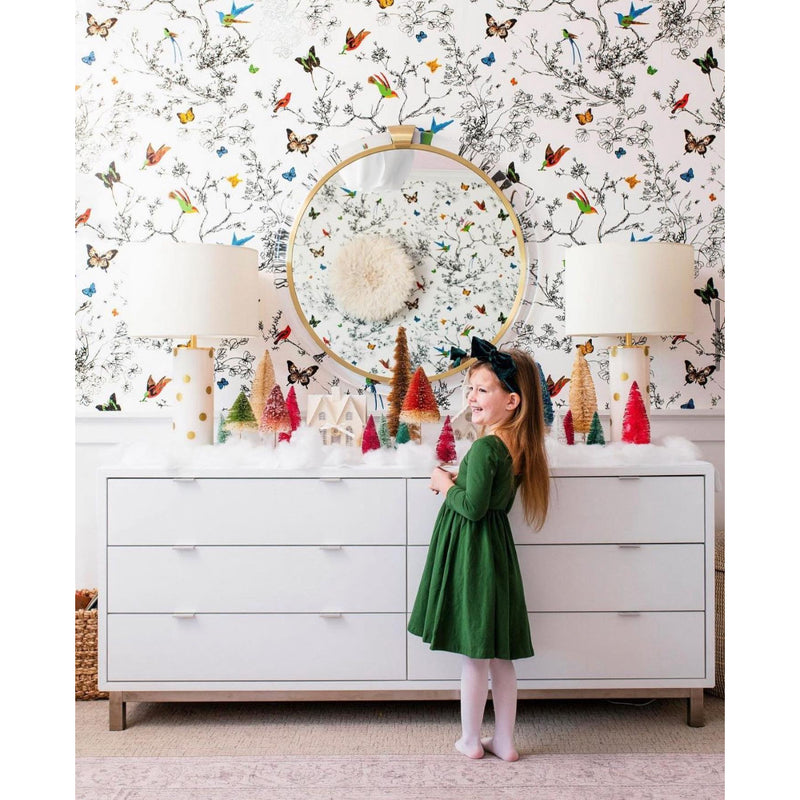 A child in a green dress stands by a decorated dresser with a polished nickel finish, featuring the Round Acrylic Mirror by Mirror Home and butterfly wallpaper.