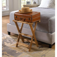 A brown leather suitcase styled like a nightstand, supported by a bamboo folding stand, placed next to a bed with a straw hat on top in a bedroom setting.