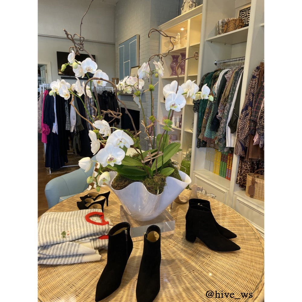 White ceramic bowl with ruffled edge filled wiht orchids on a table with shoes and clothes in the background